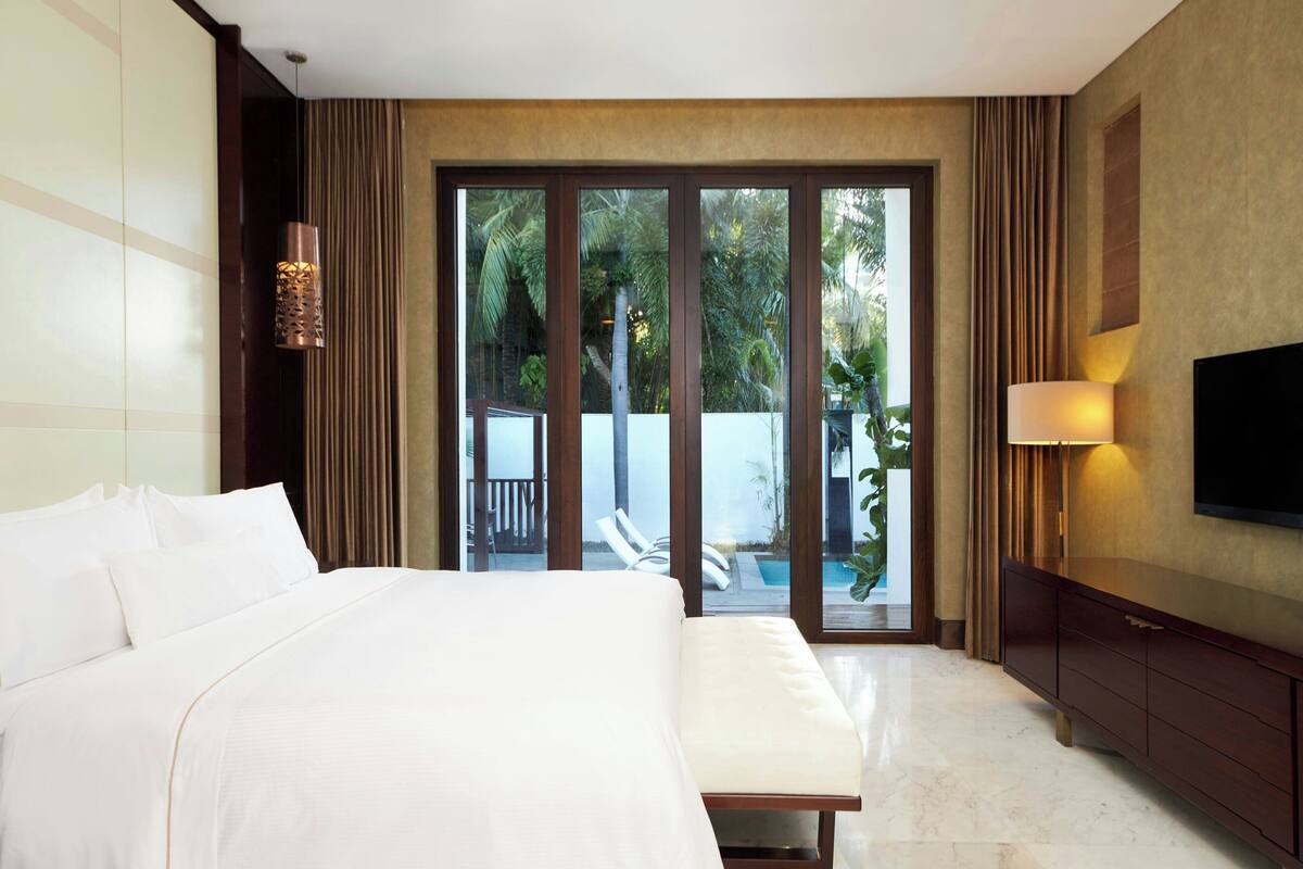 Une chambre d’hôtel avec un lit, une télévision, un banc et une vue sur une piscine et un espace verdoyant.