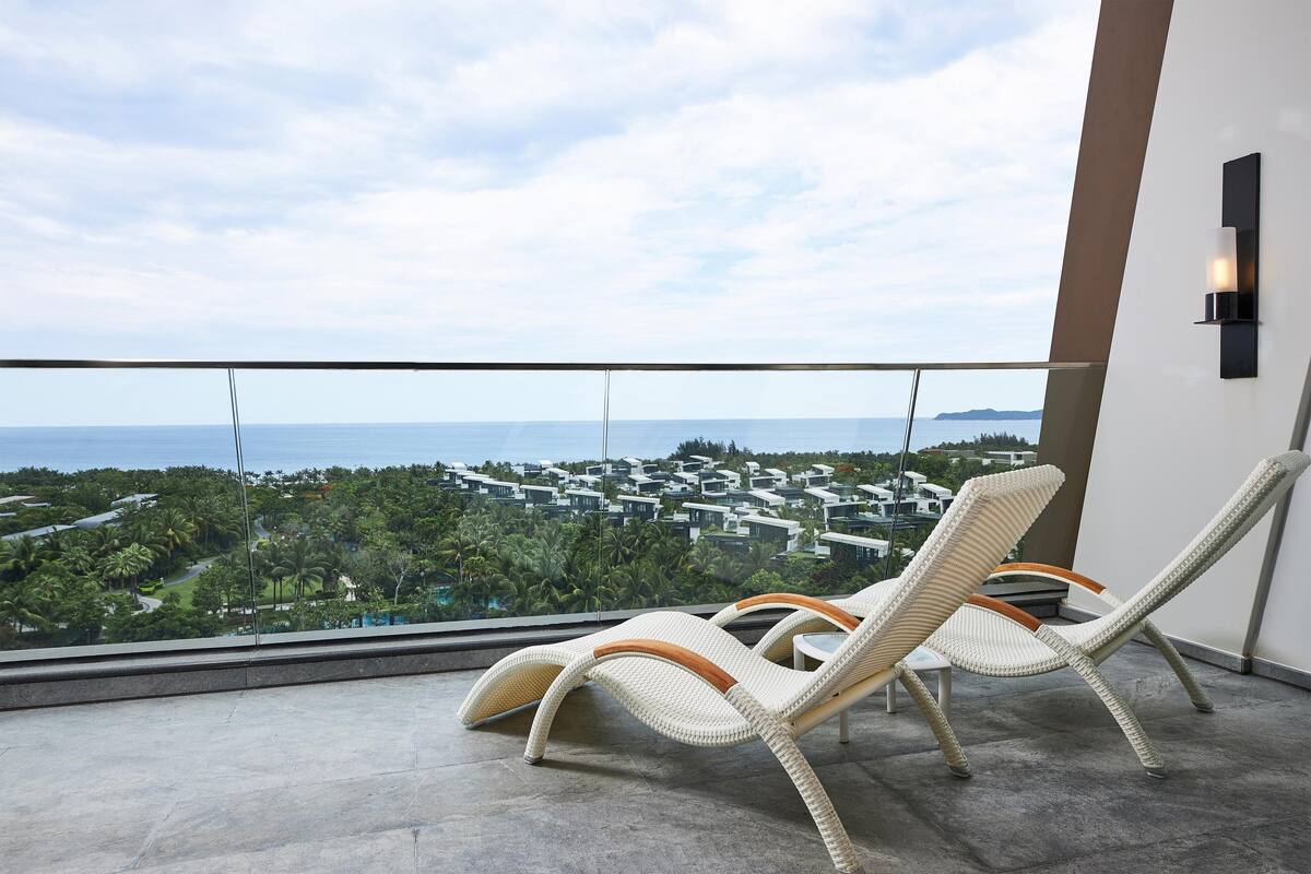 Un balcon doté de deux fauteuils de relaxation en osier, d’une balustrade en verre et offrant une vue sur une ville côtière.