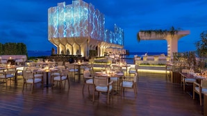 A modern building with a unique facade illuminated by blue lights, outdoor dining area with tables and chairs, and a view of the sea.