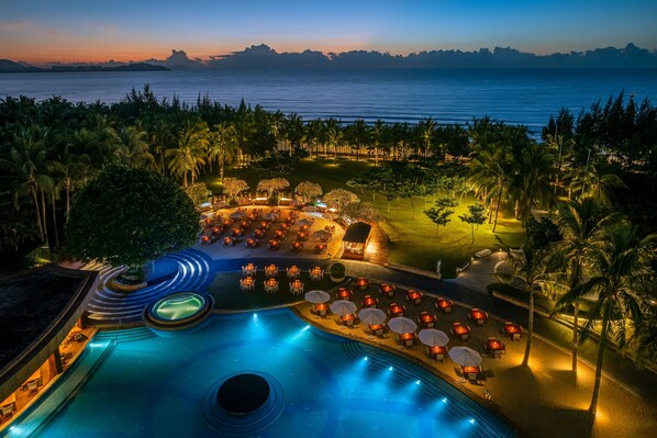 A resort pool area with a view of the ocean at dusk, surrounded by palm trees and outdoor seating.
