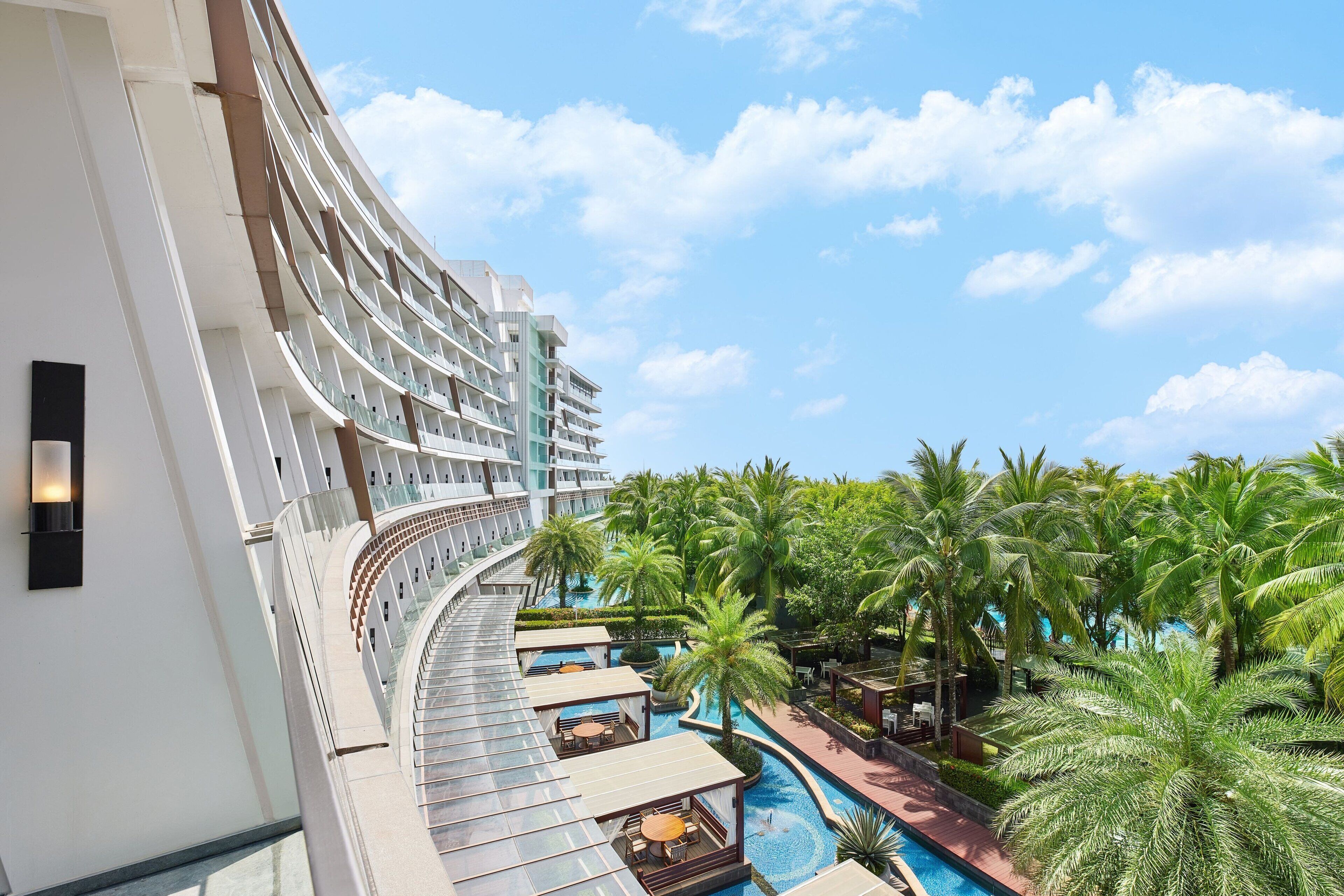 Un hôtel doté d’un balcon incurvé donnant sur un espace piscine agrémenté de palmiers et offrant une vue sur un ciel dégagé.