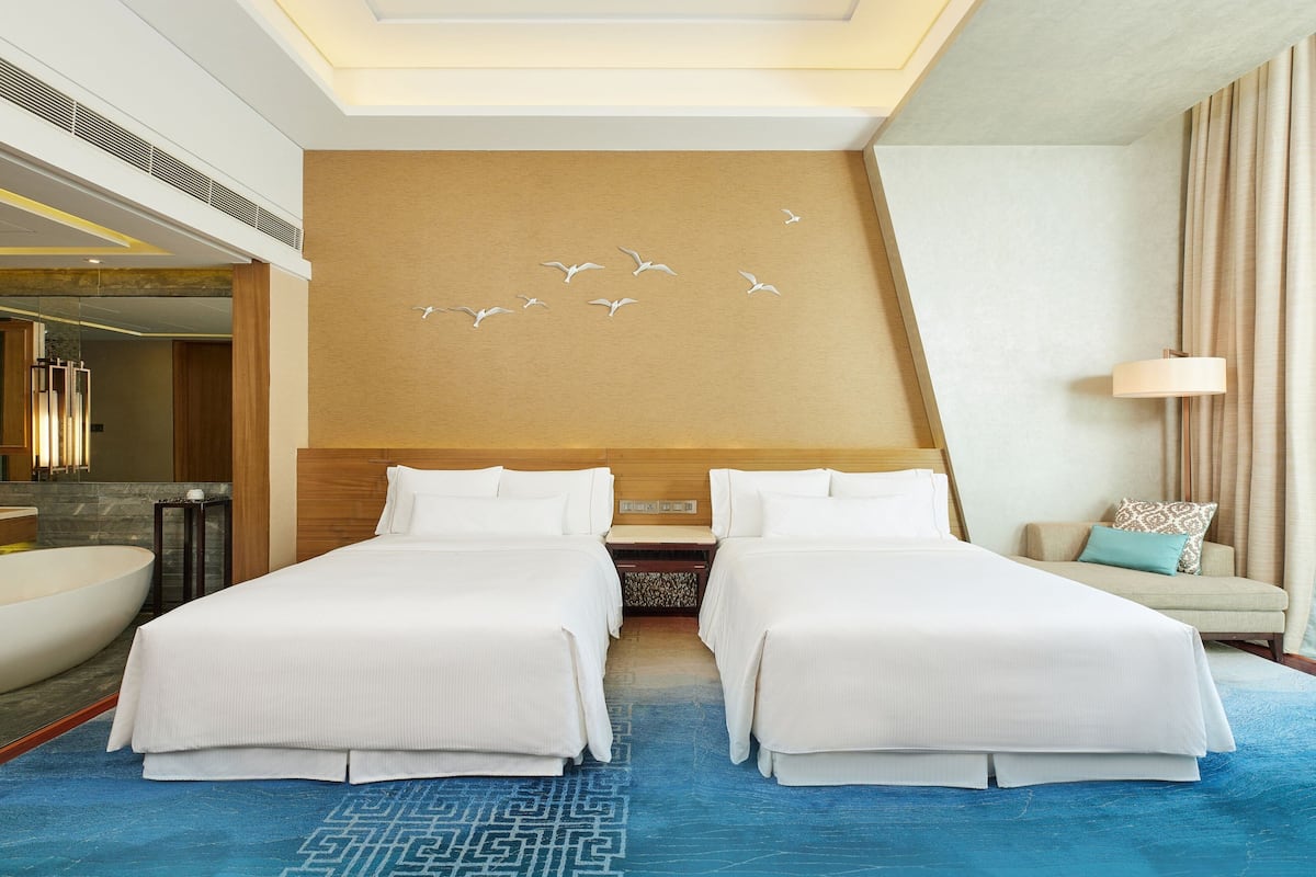 Une chambre d’hôtel avec deux lits, une grande fenêtre, une salle de bain avec baignoire et un mur décoré avec un motif d’oiseaux.