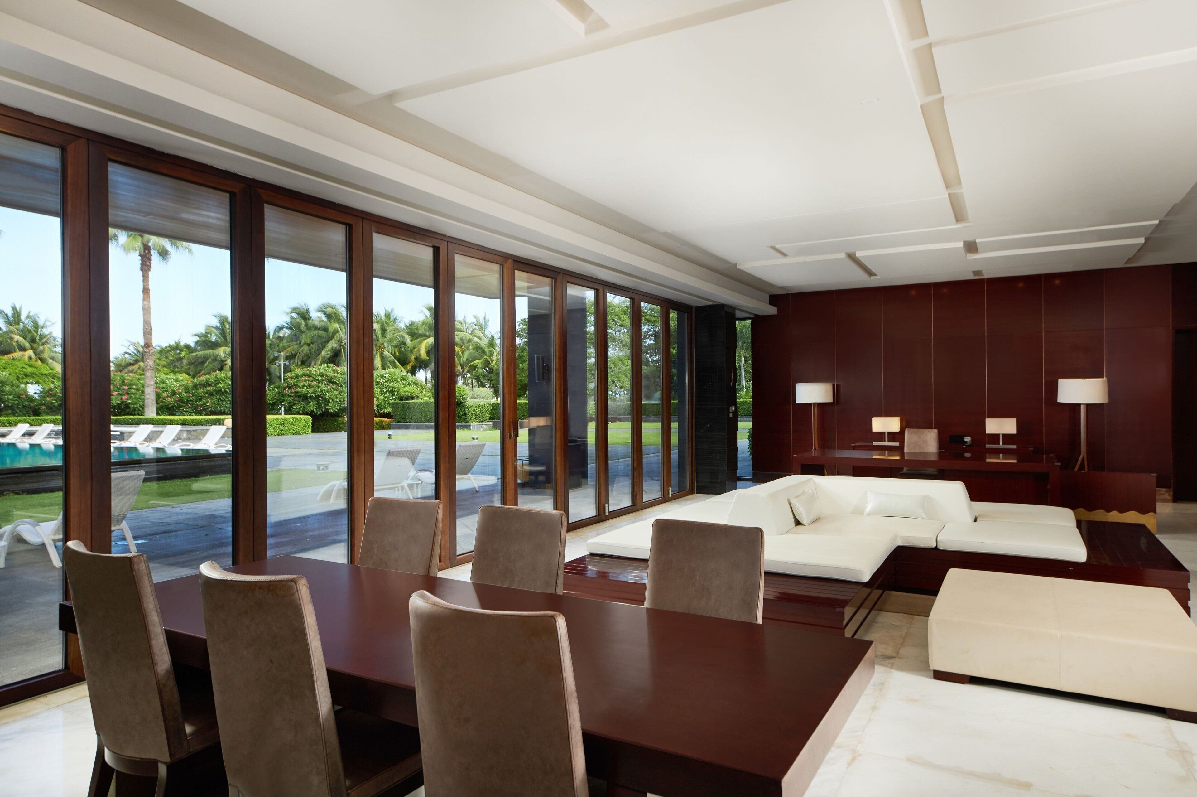 A modern hotel room with a large dining table, a white sofa, and a view of a pool and greenery.