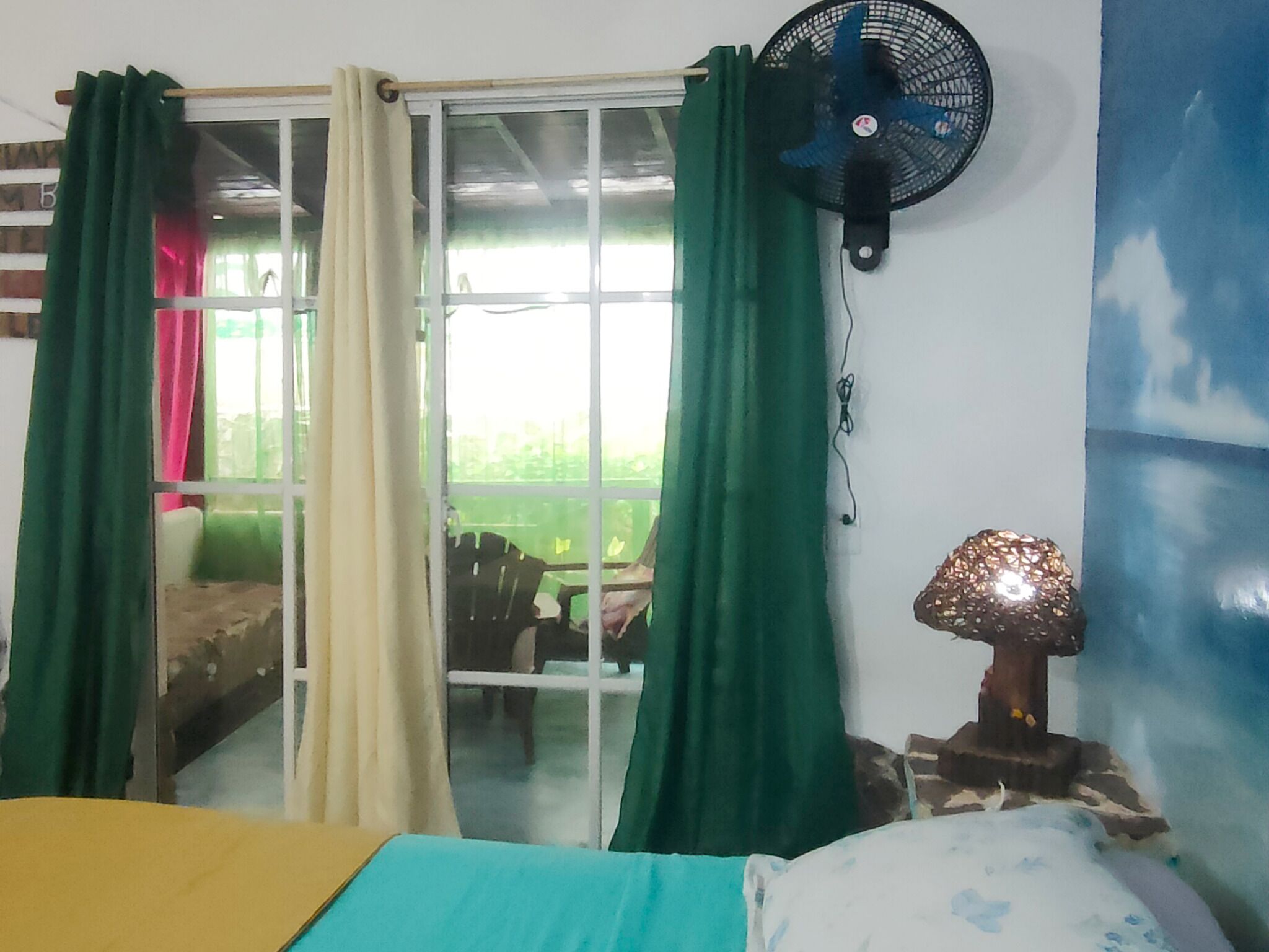 A room with a bed, a fan, a table lamp, and a window with curtains.