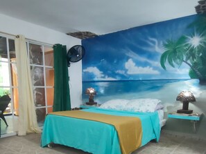 A bedroom with a bed, a nightstand, a wall-mounted fan, and a large tropical mural. - House Jardin Del Caribe (Las Terrenas)