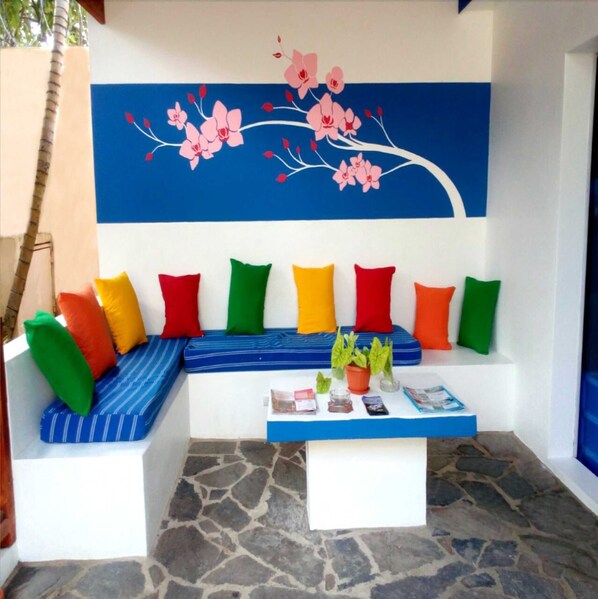 A room with a stone floor, a blue sofa with colorful cushions, a white coffee table with a plant, and a wall with a floral mural. - House Jardin Del Caribe (Las Terrenas)