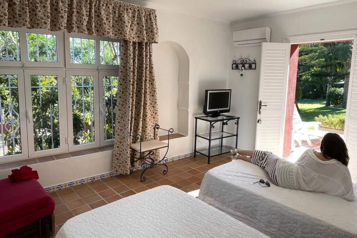 A room with two beds, a TV on a small table, and a view of a garden through an open door.