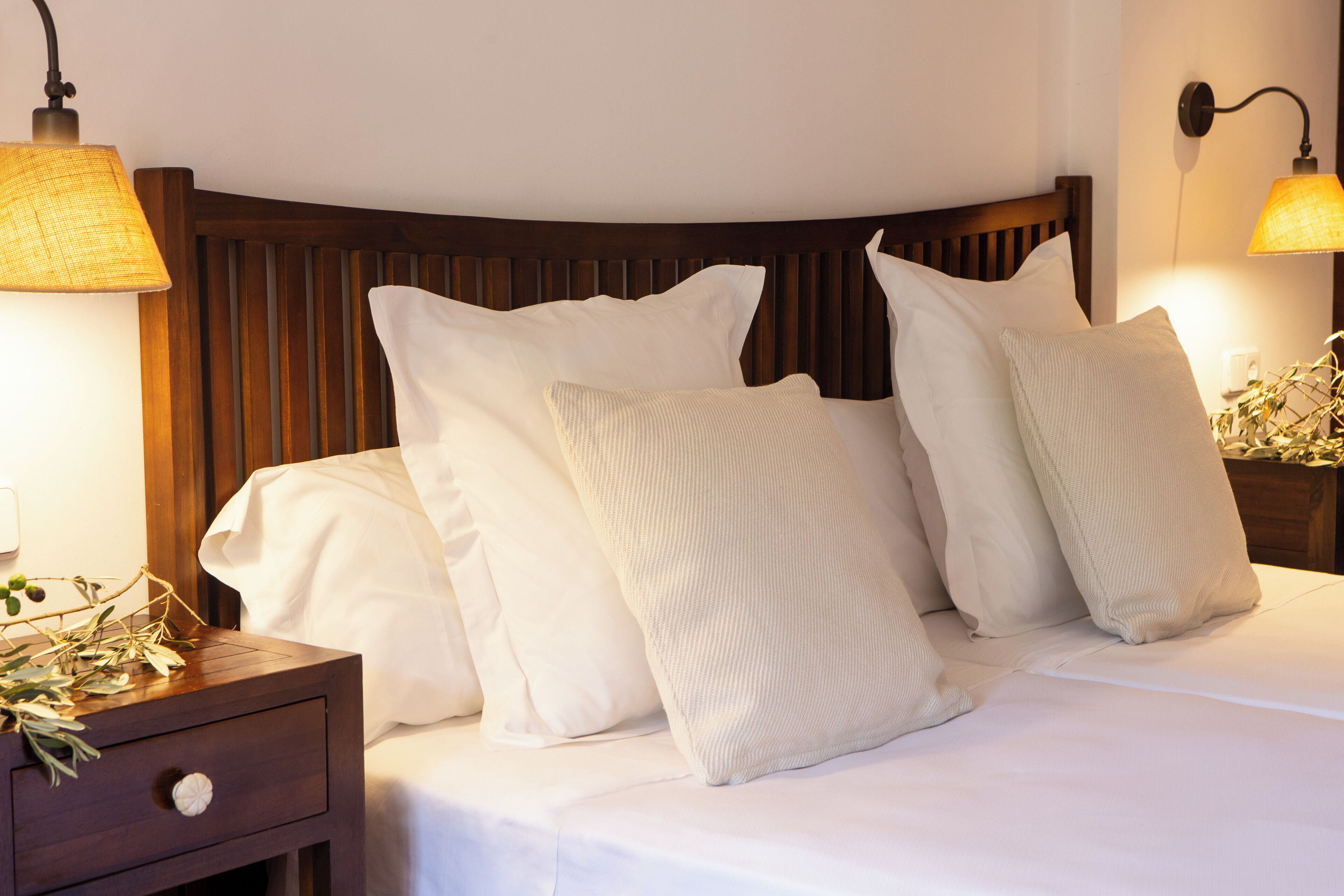 A neatly made bed with white linens and multiple pillows, a wooden headboard, and two wall-mounted lamps.