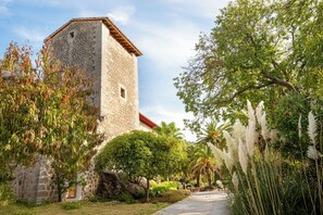 A stone building with a red-tiled roof, surrounded by lush greenery and a paved pathway.