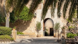 A stone building entrance with steps leading up to an arched doorway, surrounded by potted plants and palm trees. - Hotel Es Port (Sóller)