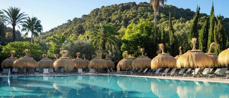 A pool area with lounge chairs and thatched umbrellas, surrounded by lush greenery and a hill in the background.