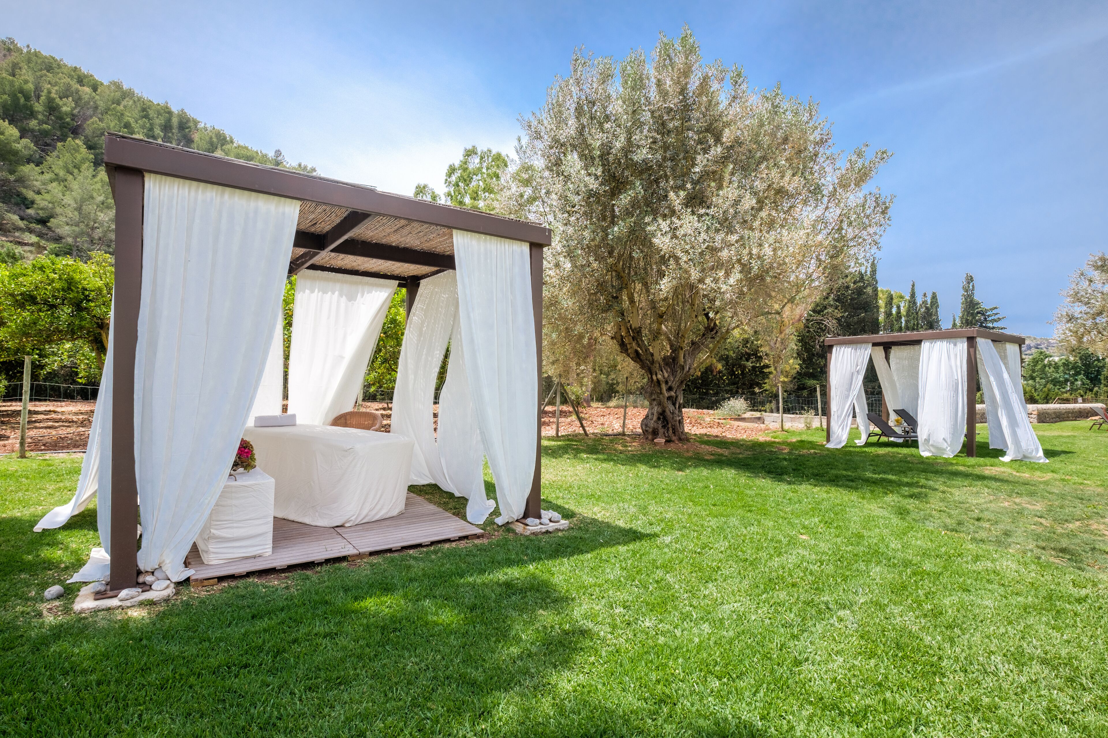 A gazebo with white drapes, a wooden bench, and a small table with a vase of flowers.