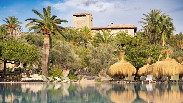 A pool with lounge chairs and thatched umbrellas, surrounded by palm trees and a stone building. - Hotel Es Port (Sóller)