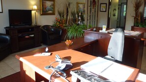 A well-lit office with a desk, chair, and television. There are plants and framed pictures on the walls.