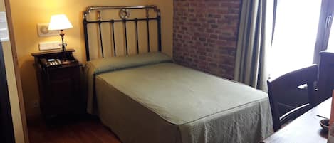 A small, single-bed room with a wooden floor, a metal headboard, a bedside table with a lamp, and a brick wall.
