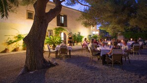 Un área de comedor al aire libre con mesas y sillas, rodeada por un árbol grande y plantas en macetas.