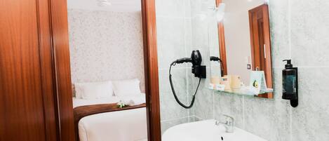 Casa de banho de hotel com porta de madeira, lavatório branco, espelho e produtos de higiene pessoal numa prateleira.