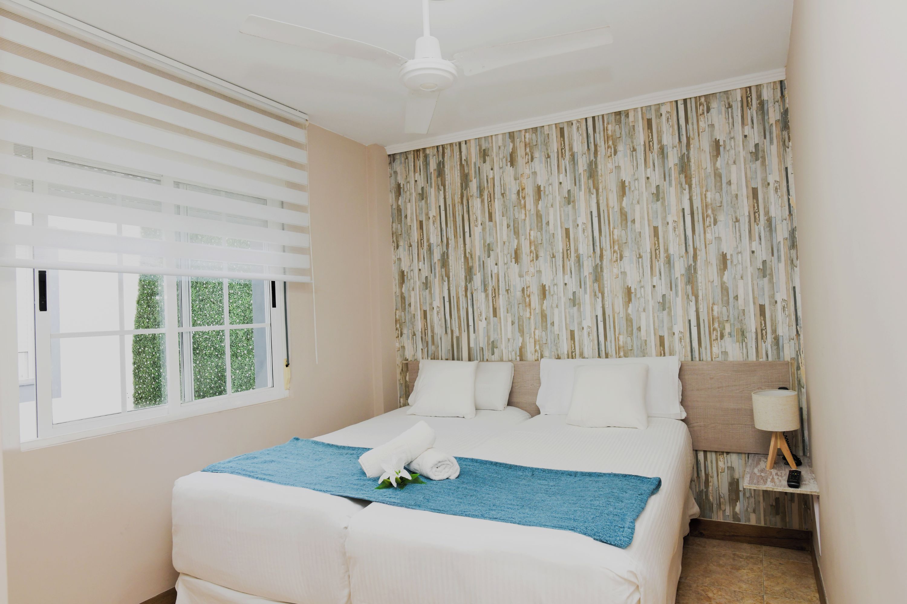 Een hotelkamer met een bed, een plafondventilator, een raam met zonwering en een wand met een verticaal patroon.