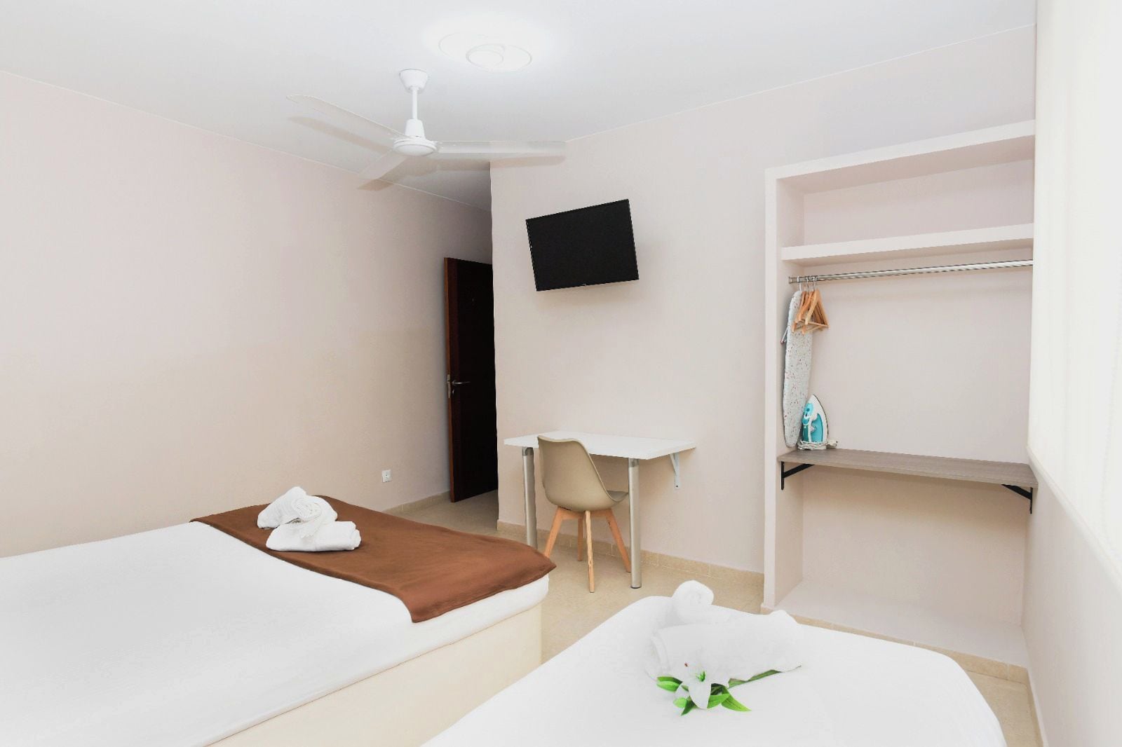 A hotel room with two beds, a desk, a chair, a TV, and a shelf with clothes and an iron.
