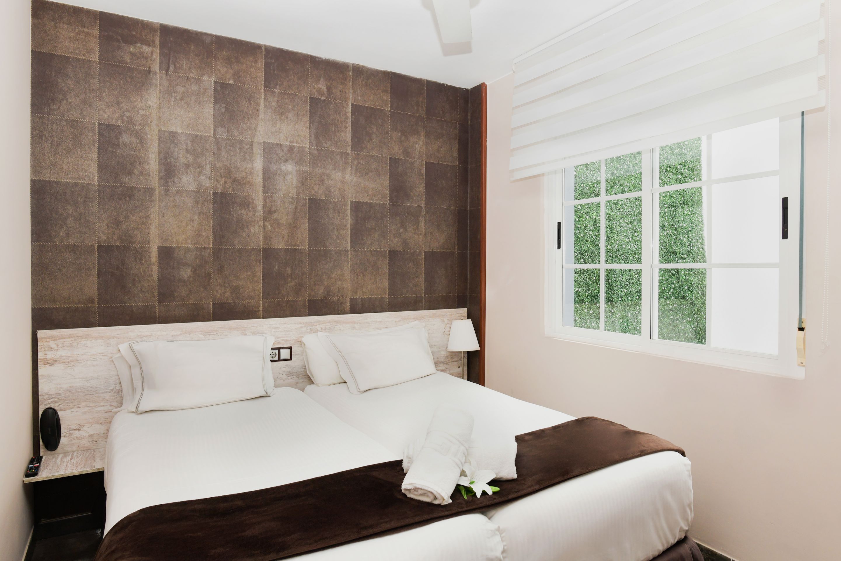 A neatly made bed with white linens and a brown throw blanket, a bedside table with a lamp, and a window with a view of greenery.