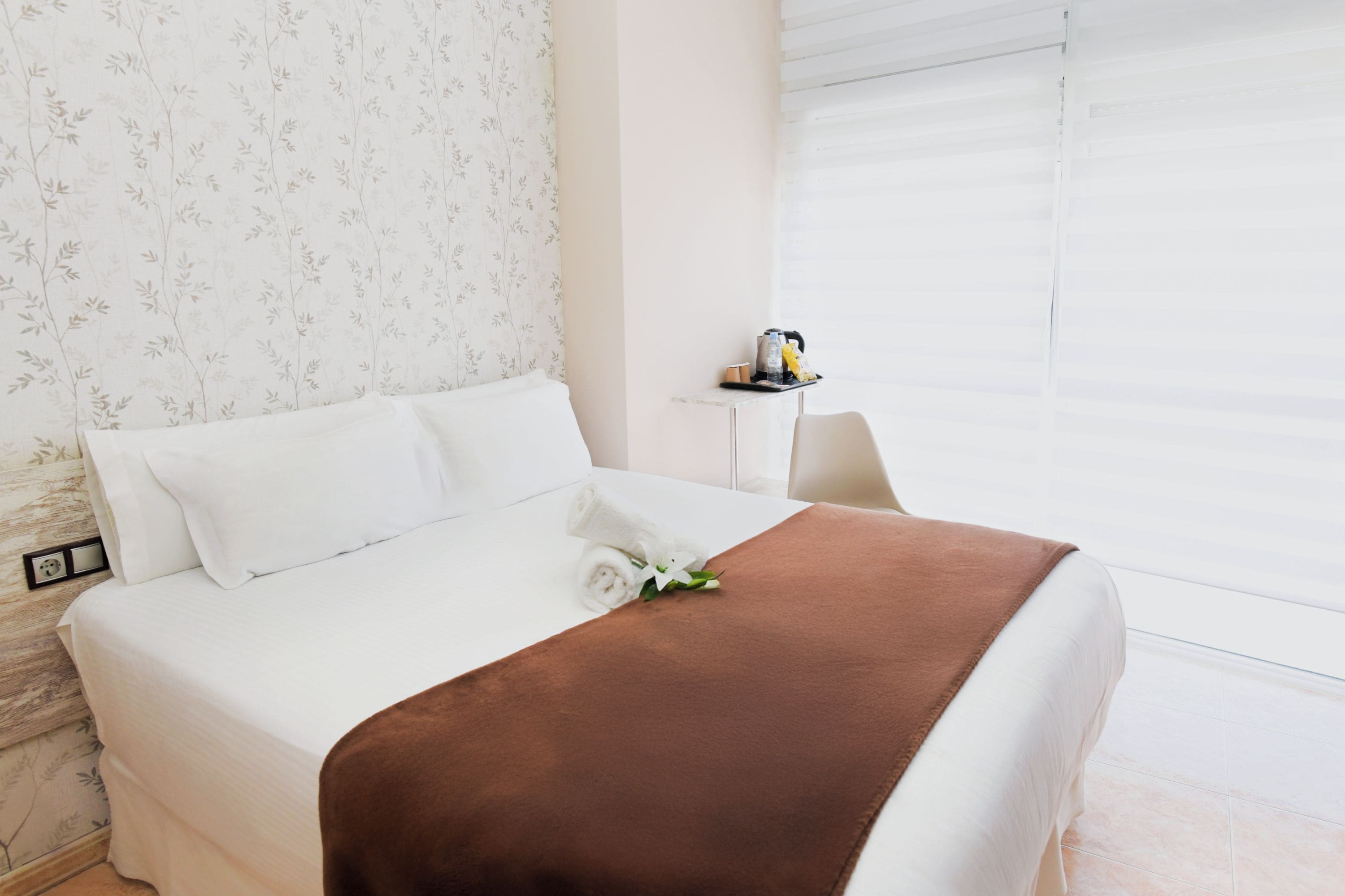 Une chambre d’hôtel avec un lit bien fait, une petite table de chevet avec une bouilloire et une tasse, et une fenêtre avec des stores.