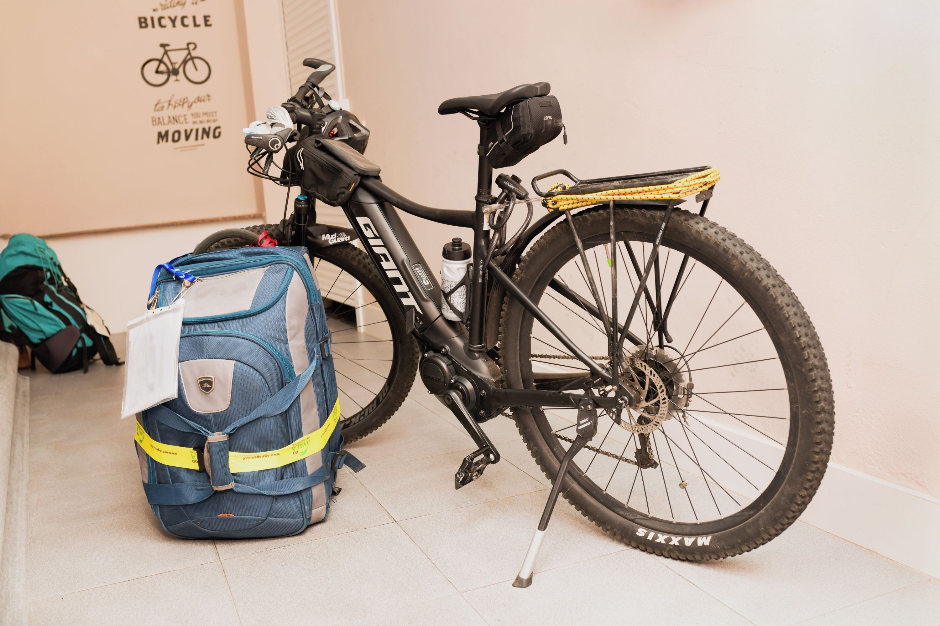 A bicycle with a black frame and a rear rack, a blue and white backpack, and a wall with a bicycle-themed poster.