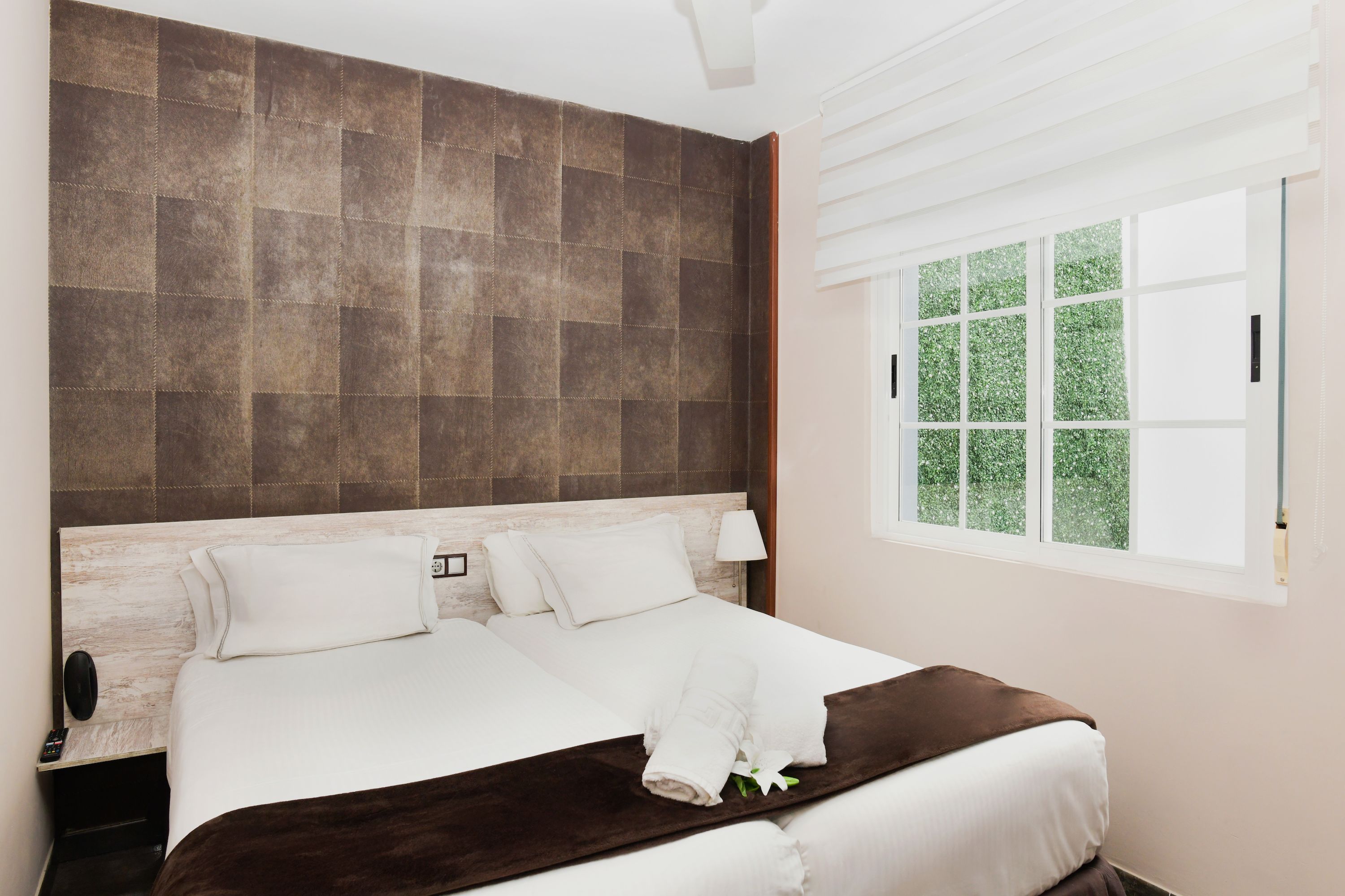 A hotel room with a large bed, a wooden headboard, a nightstand, a lamp, and a window with a view of greenery.
