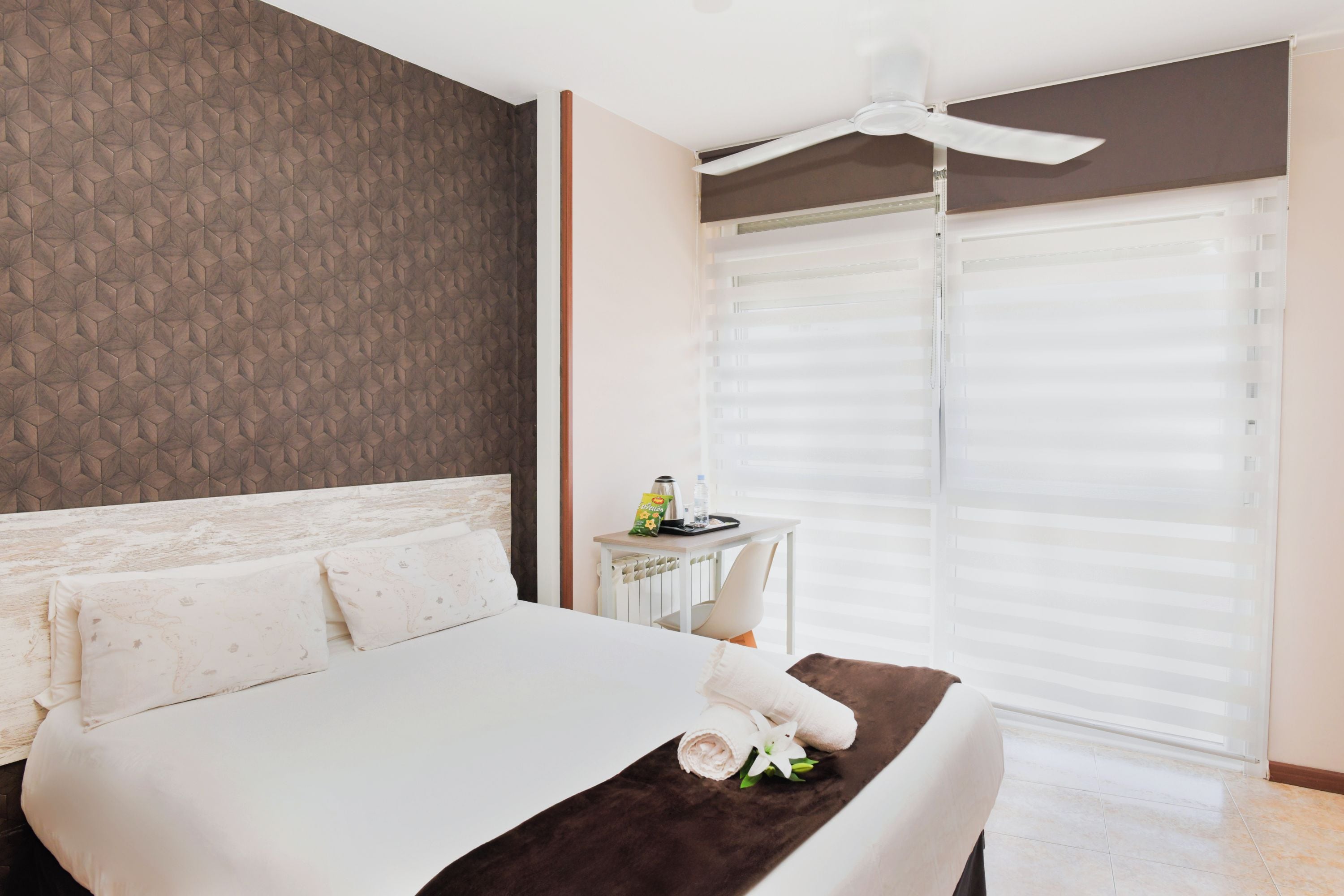 Habitación de hotel con cama, una mesita con bandeja, ventilador de techo y ventana con persianas.