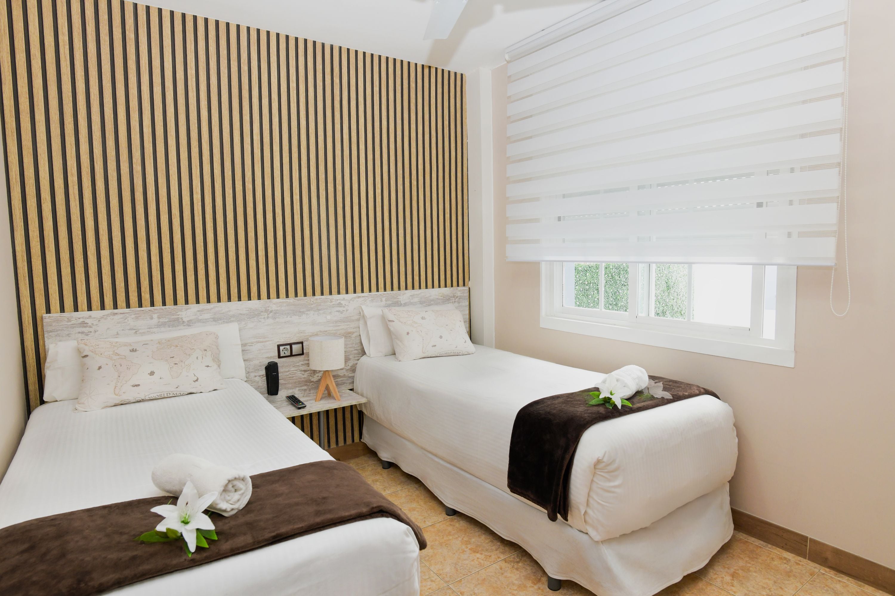 A hotel room with a wooden slat wall, two beds, a window with blinds, and a bedside table with a lamp.