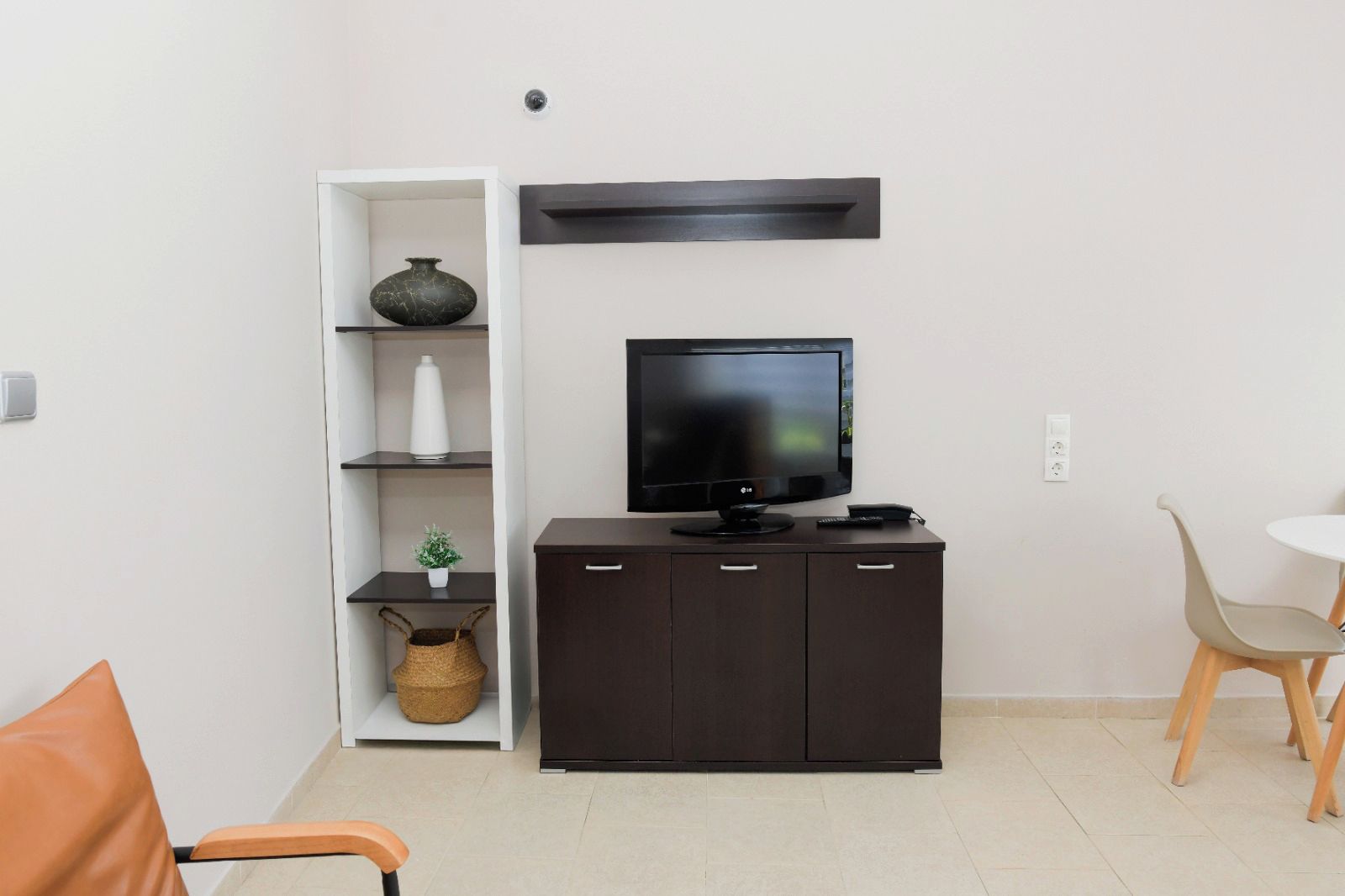 Una sala moderna con un televisor de pantalla plana, un mueble de madera oscura, una estantería blanca con objetos decorativos y un pequeño comedor con mesa y sillas.