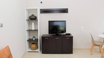 Una sala moderna con un televisor de pantalla plana, un mueble de madera oscura, una estantería blanca con objetos decorativos y un pequeño comedor con mesa y sillas.