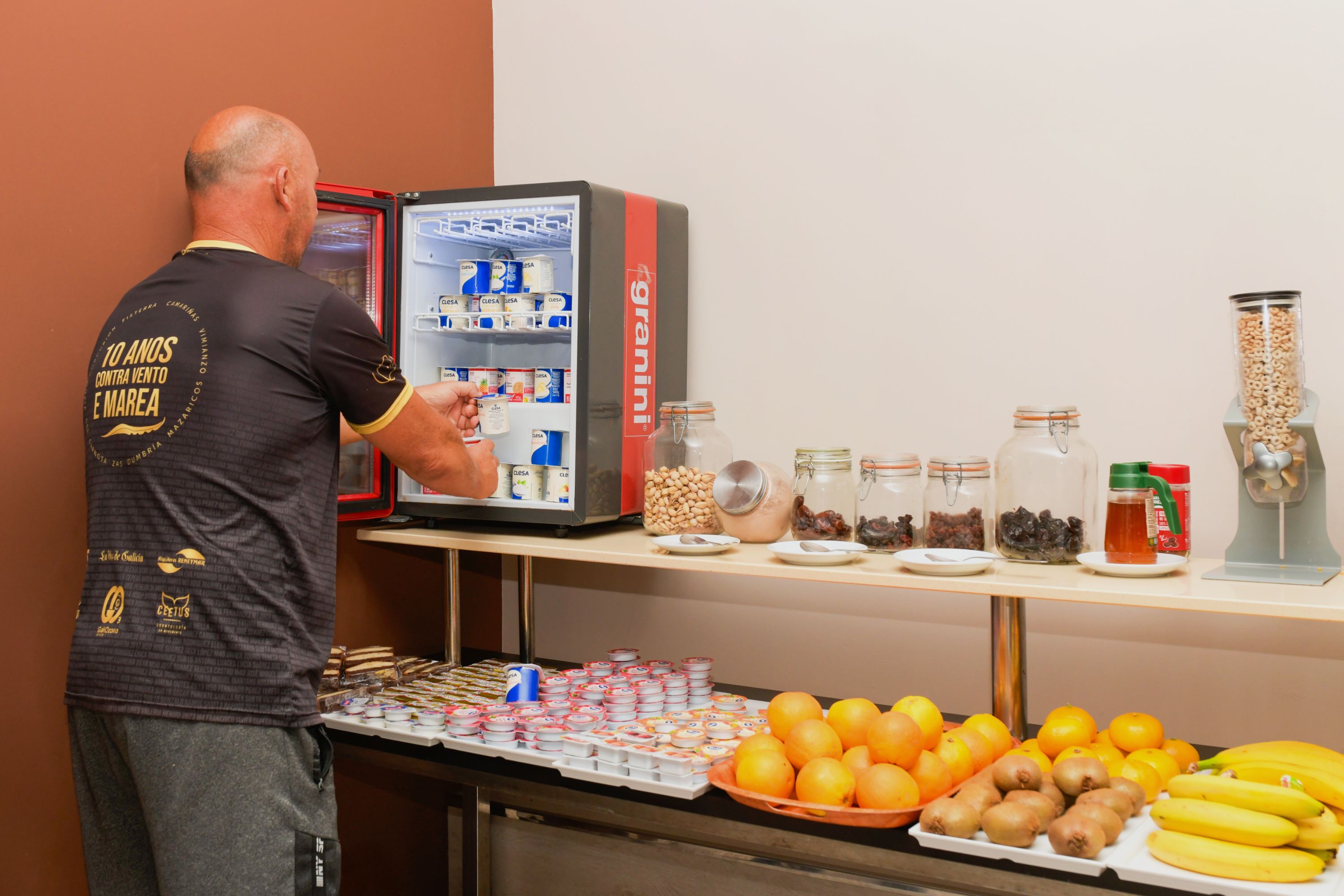 A buffet area with a refrigerator stocked with beverages, a variety of food items in jars, and a selection of fruits and snacks.