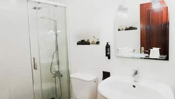 Un baño moderno con ducha envuelta en vidrio, inodoro blanco, lavamanos con espejo y un mueble de madera con artículos de aseo.