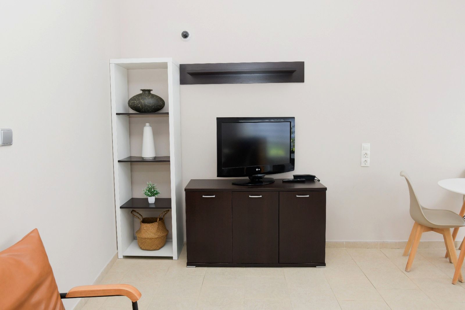 Een woonkamer met een flatscreen televisie op een donkere kast, een witte plank met decoratieve objecten, en een klein tafeltje met een stoel.