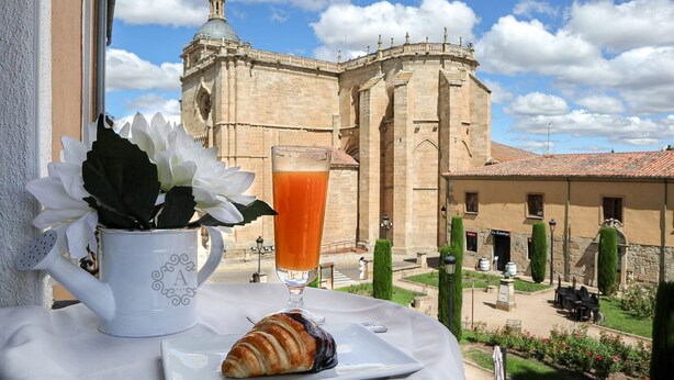 Una mesa con un croissant, un vaso de jugo de naranja y una taza blanca con una planta. Al fondo, un edificio histórico con una cúpula y un patio con sillas y una fuente.