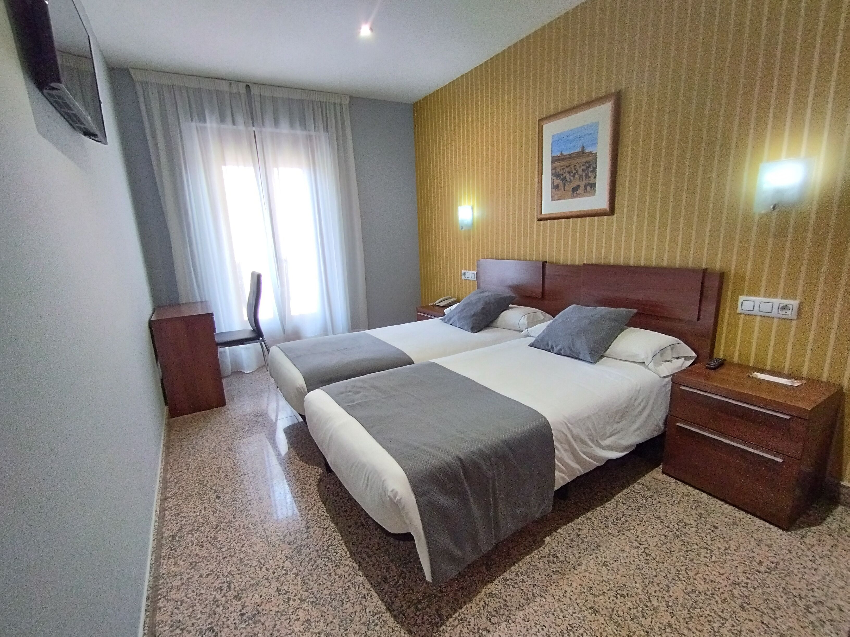 A hotel room with two beds, a desk, a chair, a nightstand, and a wall-mounted TV.