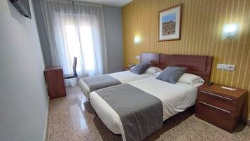 A hotel room with two beds, a desk, a chair, a nightstand, and a wall-mounted TV.