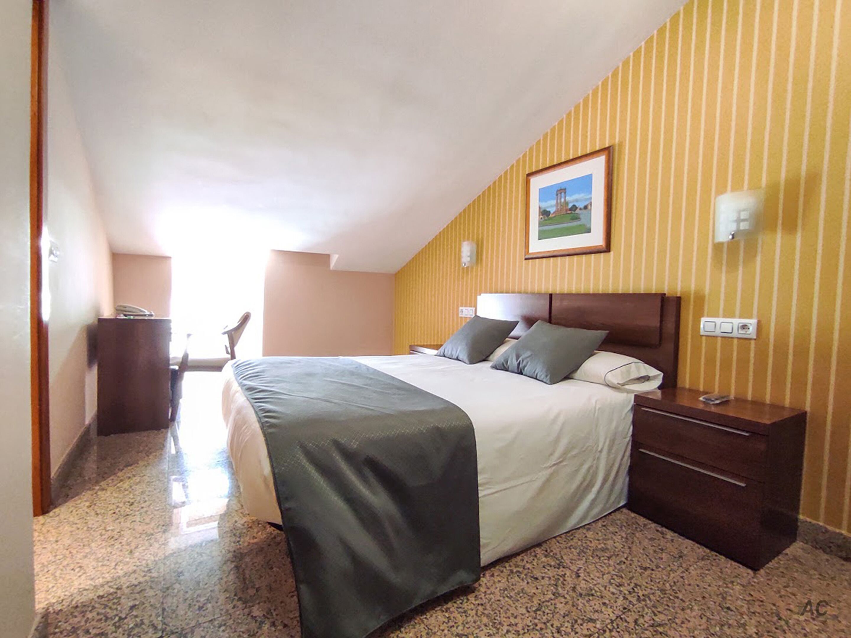 Una habitación de hotel con cama, mesita de noche, un cuadro en la pared y ventana.