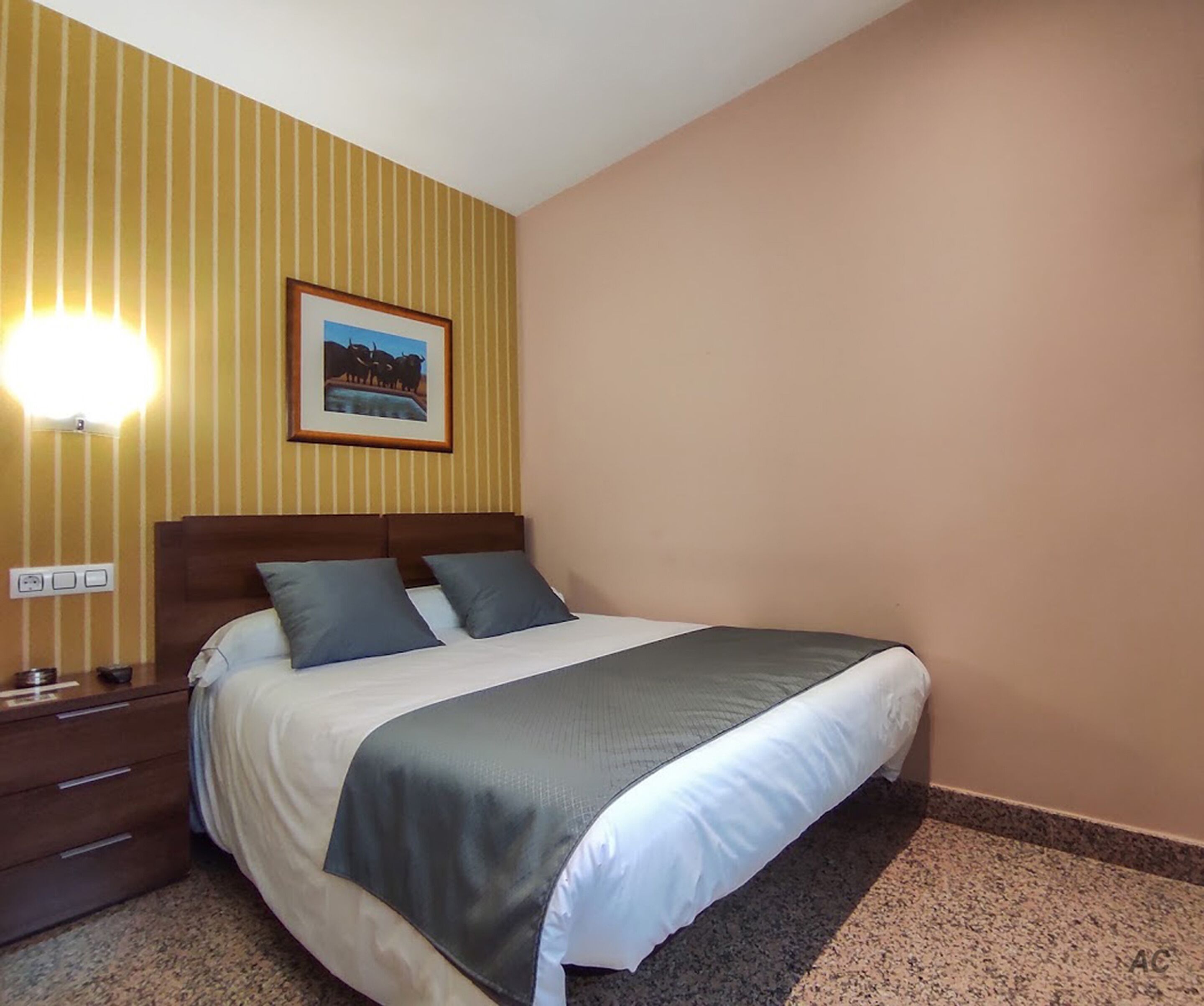 Una habitación de hotel con una cama, una mesita de noche, una lámpara de pared e imagen enmarco.