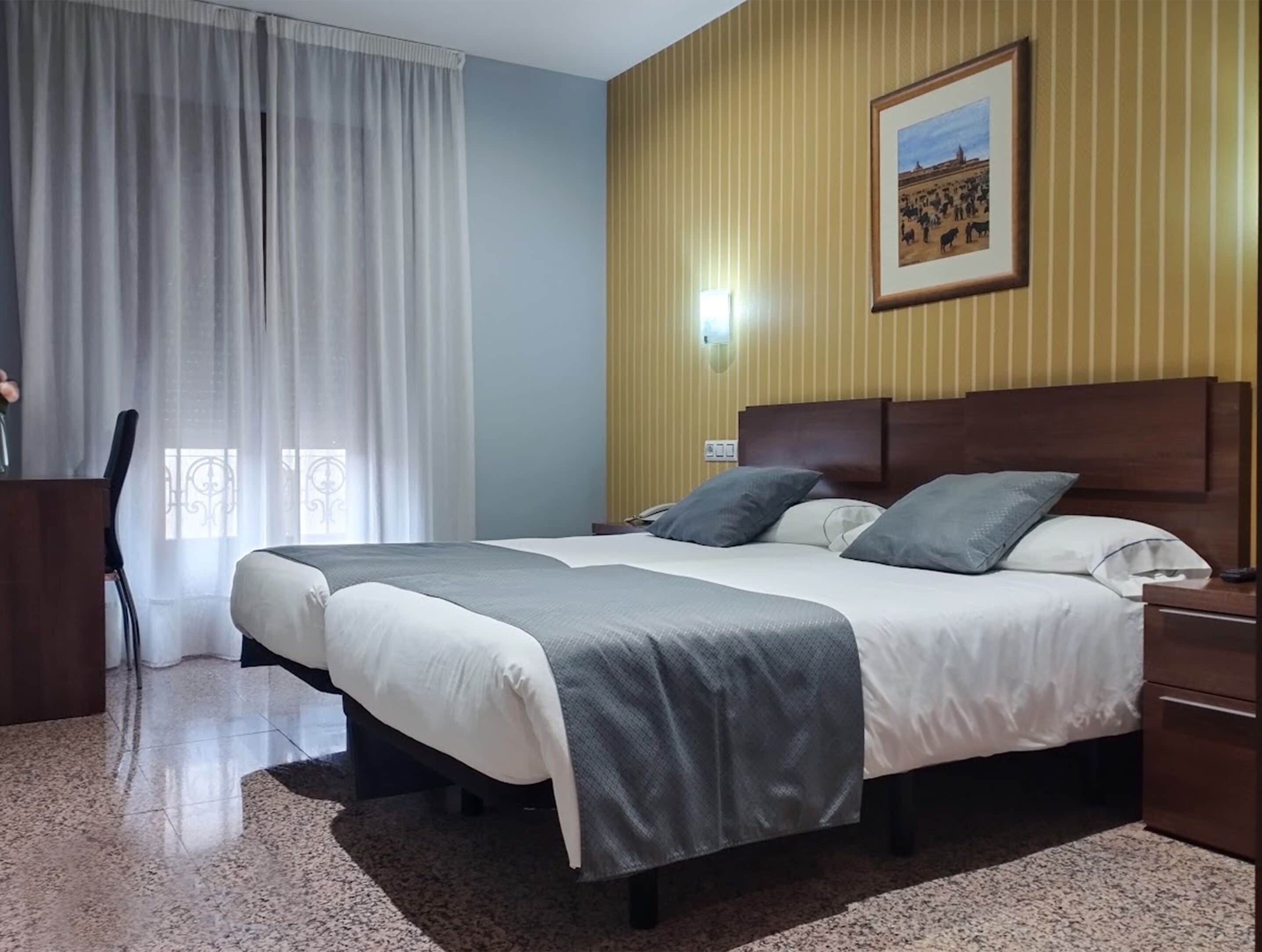 A hotel room with a bed, a desk, a chair, a window with curtains, and a wall-mounted light.