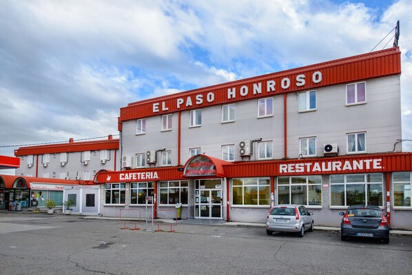A two-story building with a red roof and signage for 'El Paso Honroso Restaurante' and 'Cafeteria'. There are two cars parked in front. - El Paso Honroso (Hospital de Orbigo)