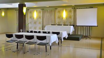 Un salón de conferencias con mesas dispuestas para una reunión, sillas, una pantalla de proyector y decoración floral en las paredes.