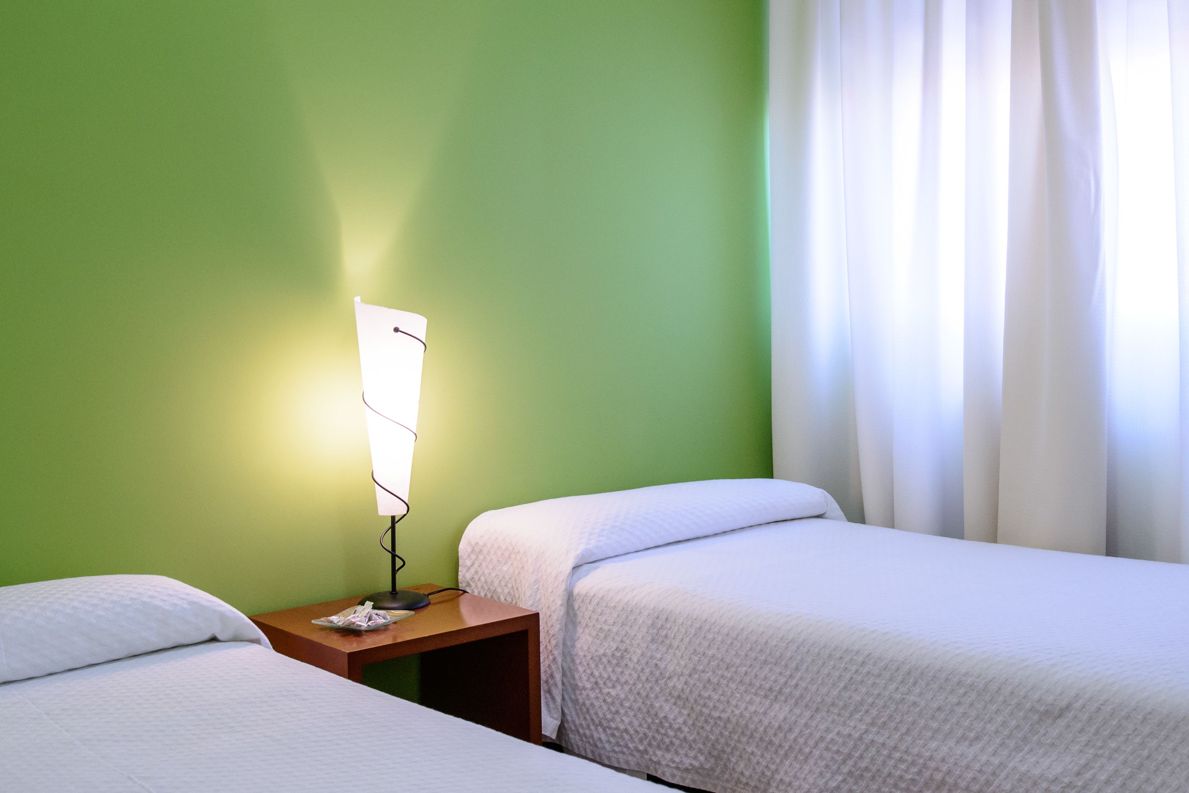 Habitación de hotel con dos camas individuales, una mesita de noche con lámpara y una ventana con cortinas.
