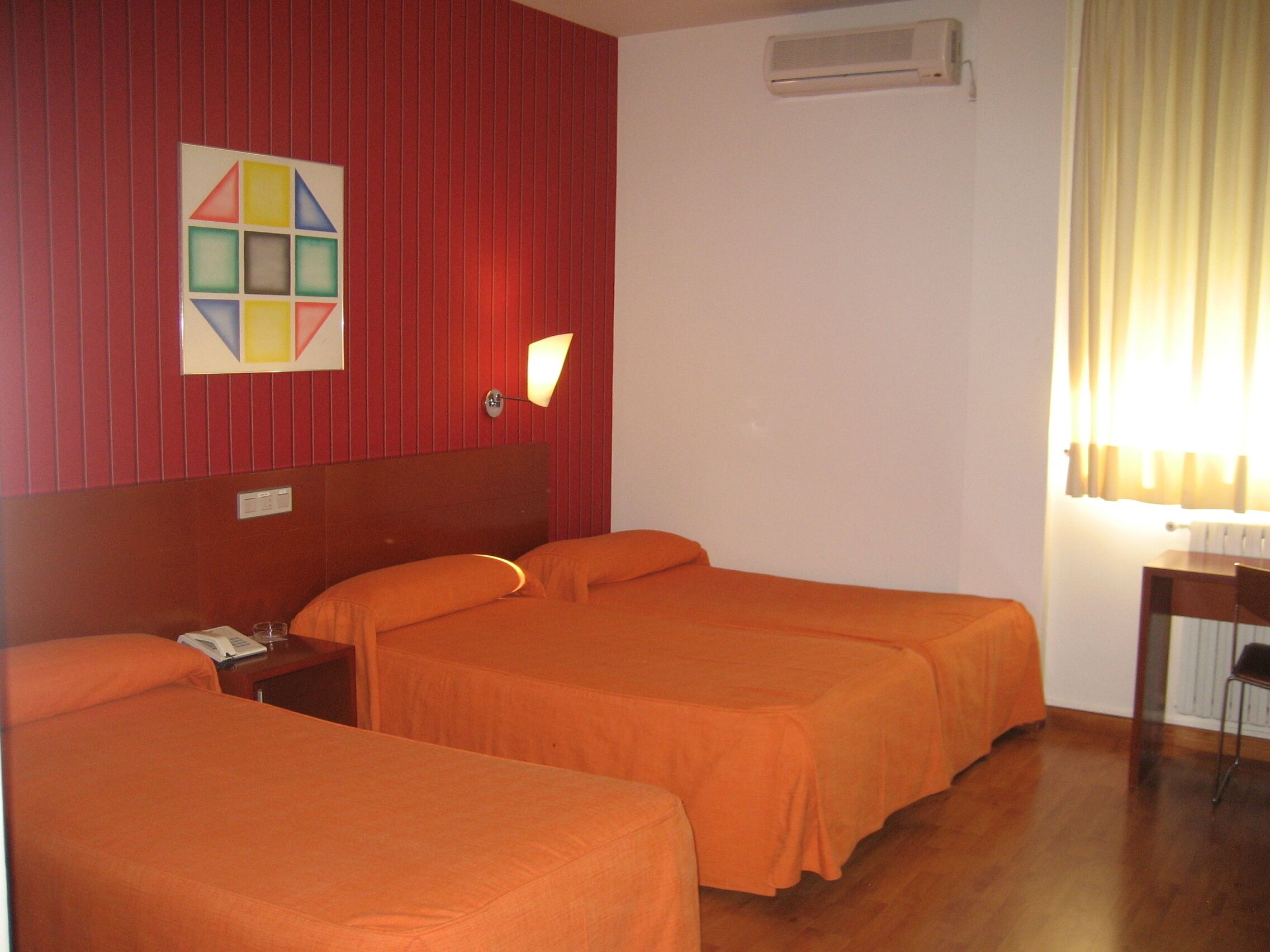 Habitación de hotel con dos camas, aire acondicionado de pared, mesita de noche con lámpara y ventana con cortinas.