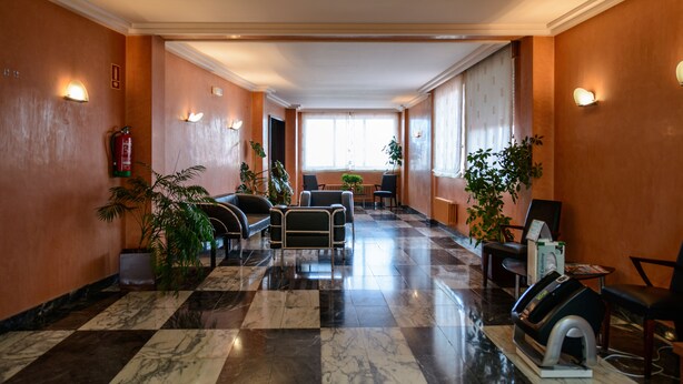 A hotel lobby with a checkered marble floor, seating area with chairs and tables, potted plants, and wall-mounted light fixtures. - El Paso Honroso (Hospital de Orbigo)