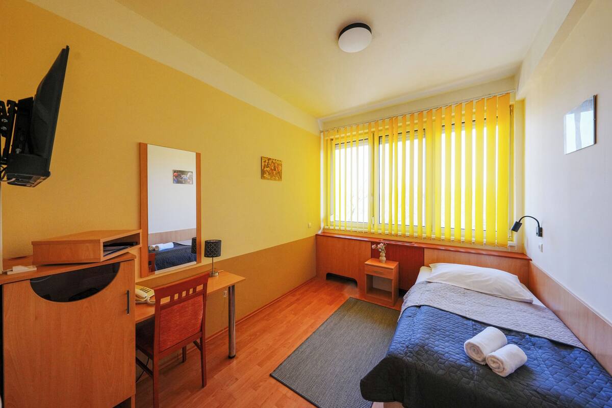 A hotel room with a bed, desk, chair, and a window with blinds.