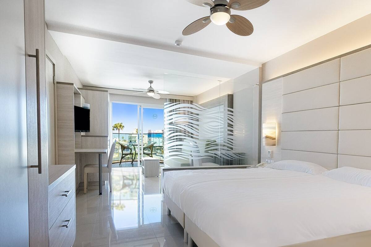 A modern hotel room with a large bed, a ceiling fan, a TV, and a balcony with a view of the city.
