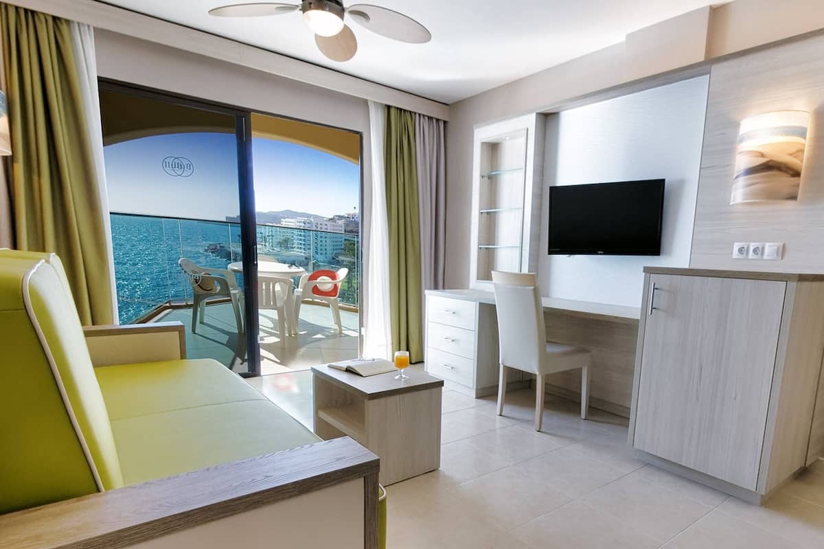 A hotel room with a balcony, a desk, a chair, a TV, and a minibar.