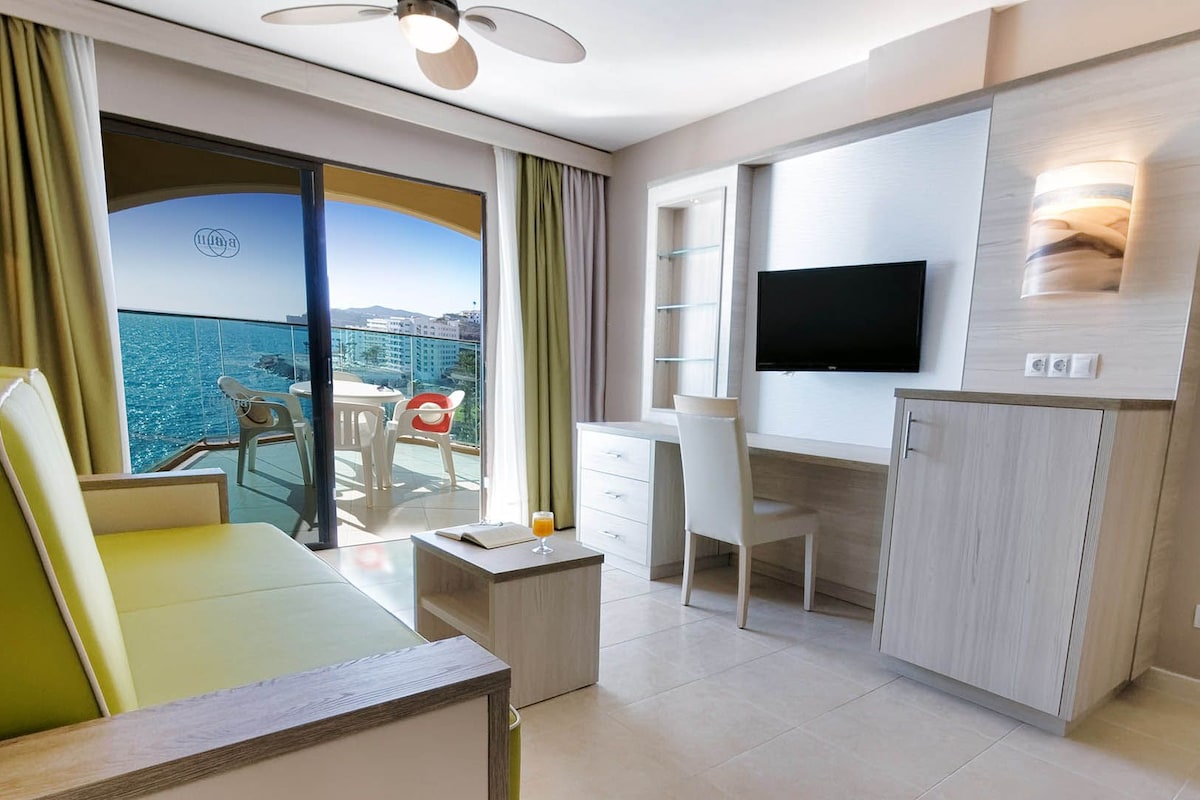 A hotel room with a balcony, a desk, a chair, a television, and a wardrobe.