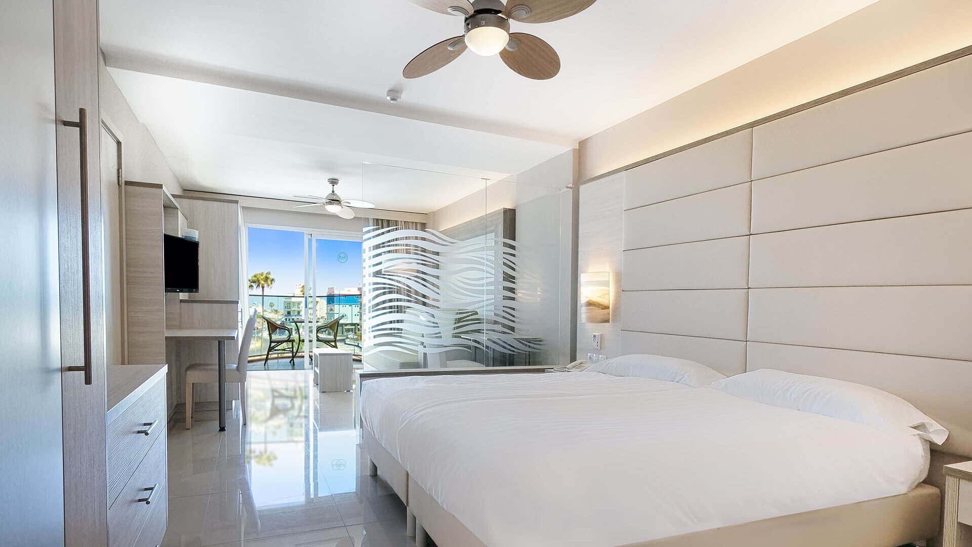 A modern hotel room with a large bed, a ceiling fan, a TV, and a balcony with a view of the city.
